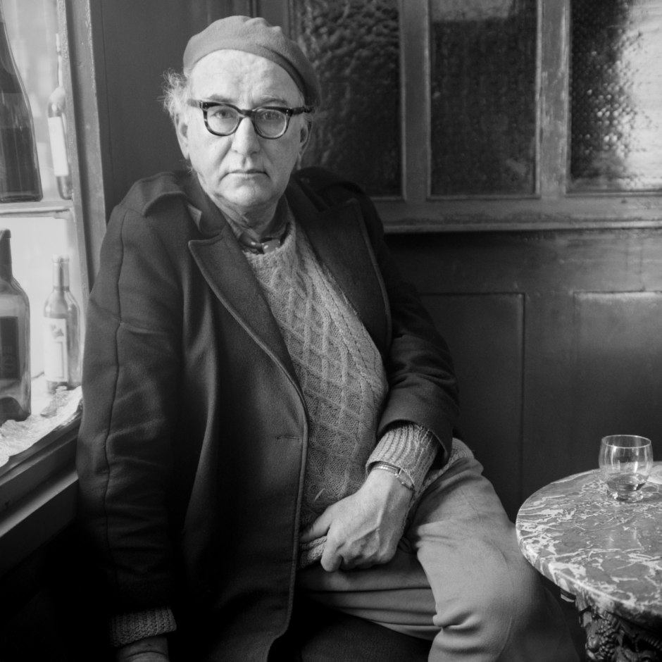 Black and white photo portrait of poet Patrick Kavanagh. He is seated, legs crossed, by a window next to a small table